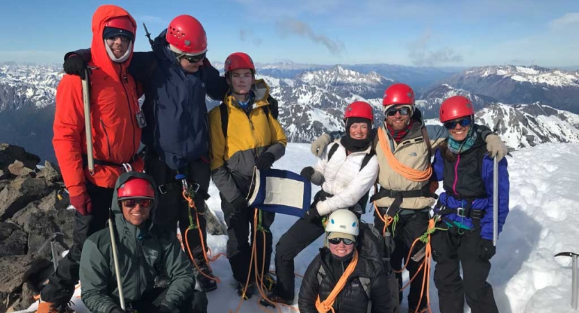 A group of people wearing safety and cold-weather gear stand on a snowy outlook and pose for a photo. Two people are holding a blue peter flag. In the background, there is a snowy, mountainous landscape. 